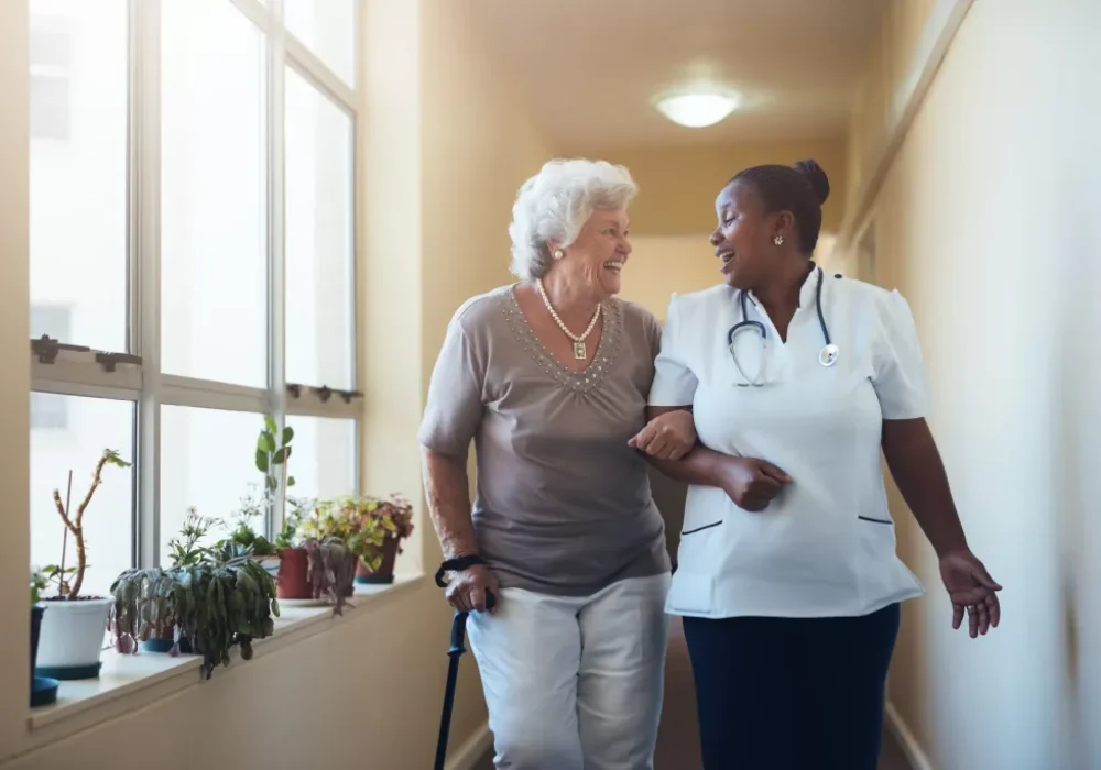 smiling-healthcare-worker-and-senior-woman-walking-2021-08-26-19-58-19-utc-scaled